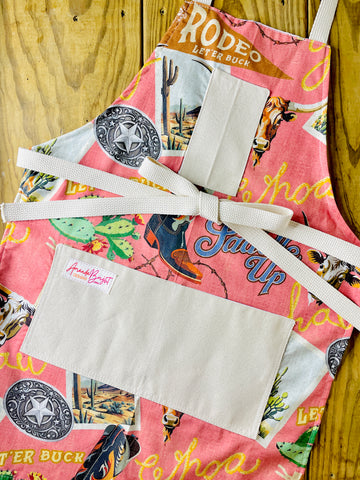 Amanda Bright Design colorful cotton apron with cowboy-themed design let ‘er buck saddle up buckle cowboy boot cactus postcard western