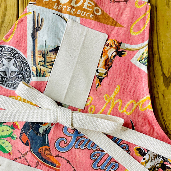 Amanda Bright Design colorful cotton apron with cowboy-themed design let ‘er buck saddle up buckle rodeo cowboy boot cactus postcard western