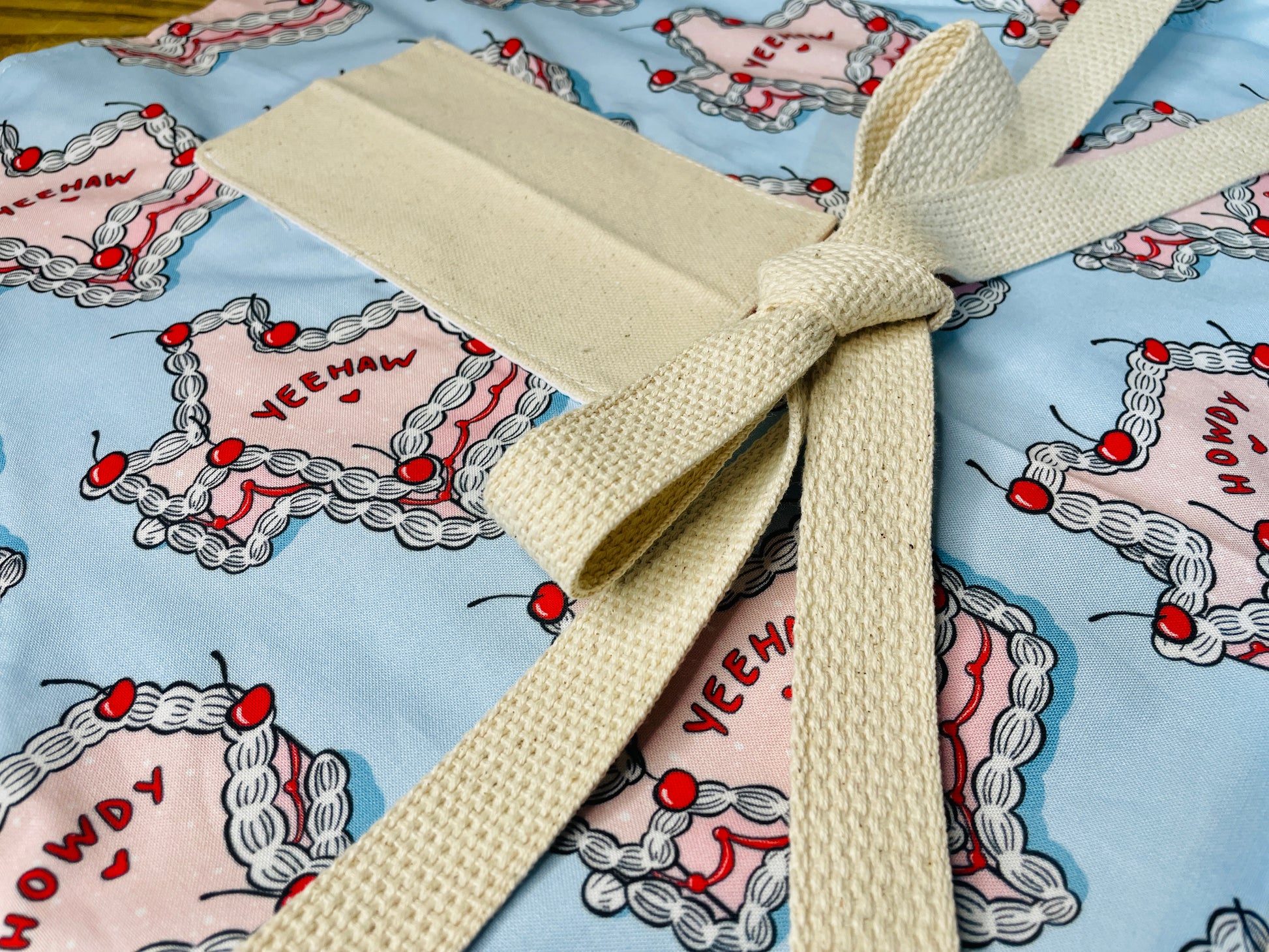 Amanda Bright Designs Texas Cake Couture Apron featuring texas shaped like a cake with pink icing cherries and words yeehaw and howdy written on the cake on a baby blue background