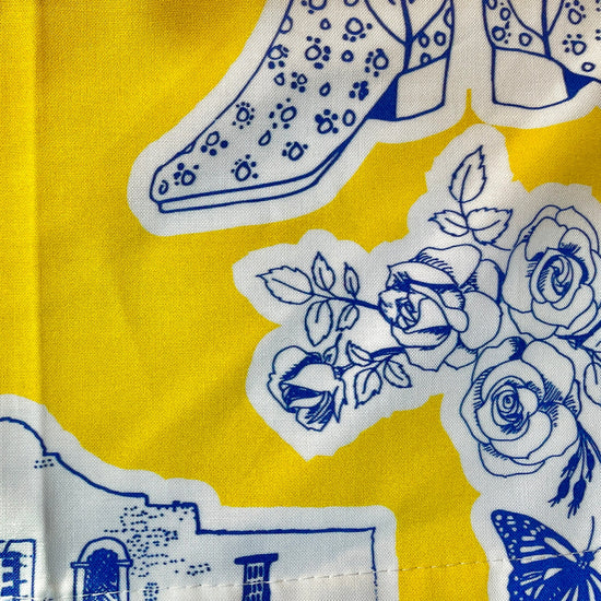 Amanda Bright Designs Texas Toile Apron feaures cowboy boots deer cactus the alamo roses in blue and white on gold background
