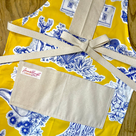 Amanda Bright Designs Texas Toile Apron feaures cowboy boots deer cactus the alamo roses in blue and white on gold background
