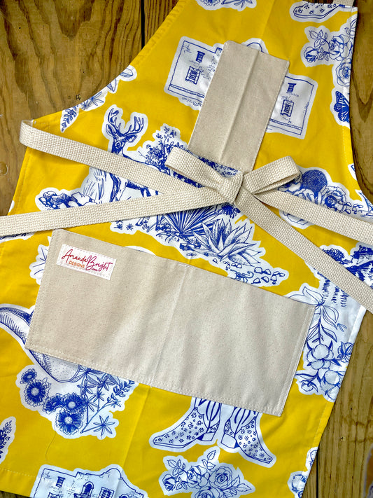Amanda Bright Designs Texas Toile Apron feaures cowboy boots deer cactus the alamo roses in blue and white on gold background
