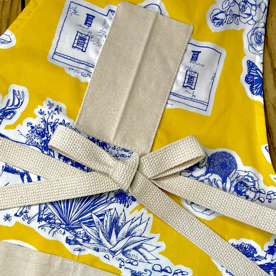 Amanda Bright Designs Texas Toile Apron feaures cowboy boots deer cactus the alamo roses in blue and white on gold background
