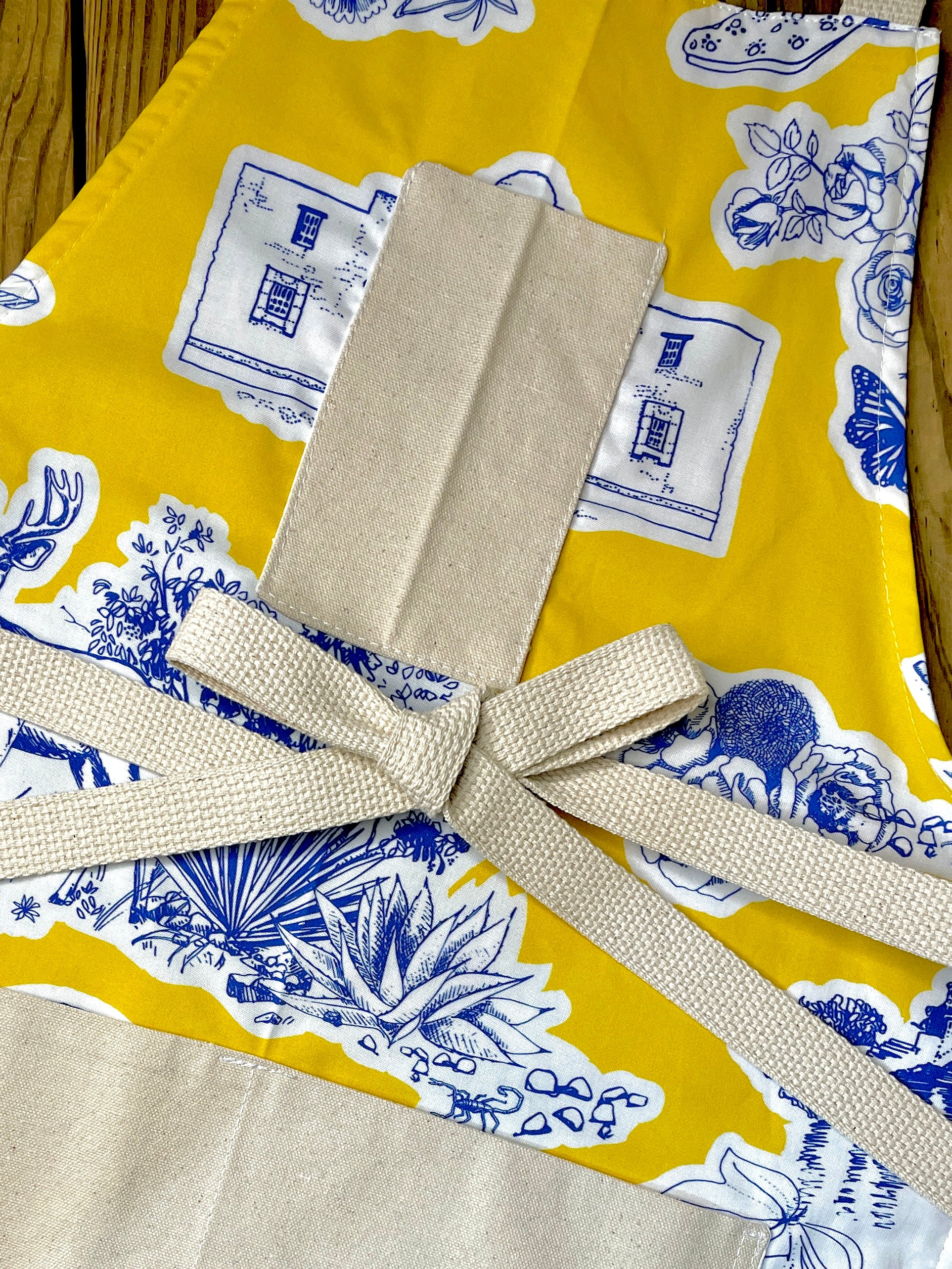 Amanda Bright Designs Texas Toile Apron feaures cowboy boots deer cactus the alamo roses in blue and white on gold background
