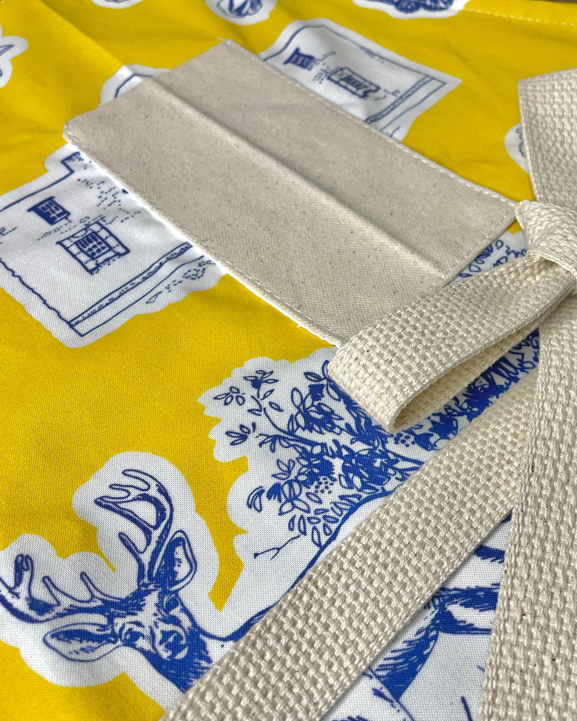 Amanda Bright Designs Texas Toile Apron feaures cowboy boots deer cactus the alamo roses in blue and white on gold background
