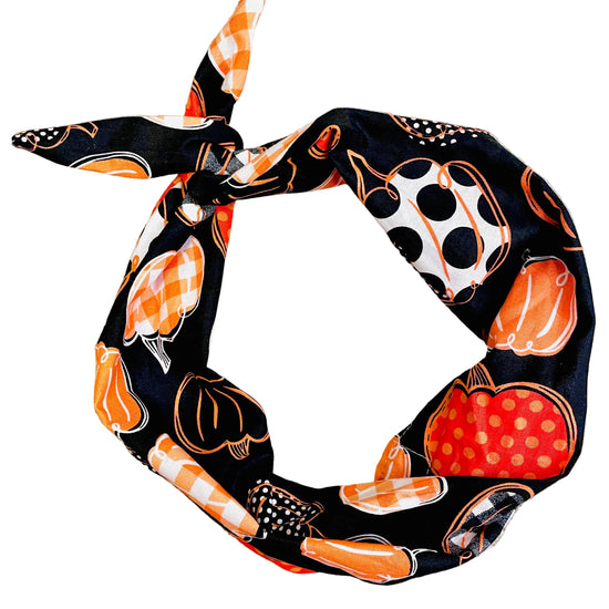 A black fabric headband with a Halloween pumpkin pattern, tied in a knot and laid flat on a beige carpet. The pattern features orange pumpkins, white pumpkins with black polka dots, and orange-and-white checkered stripes.