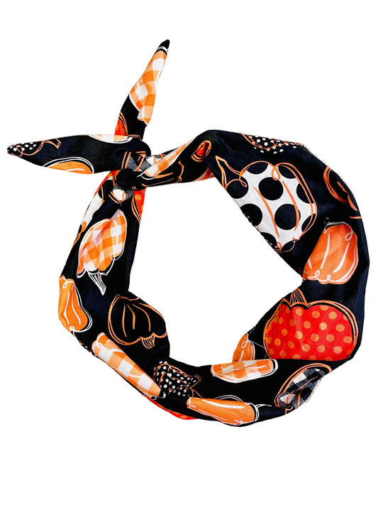 A black fabric headband with a Halloween pumpkin pattern, tied in a knot and laid flat on a beige carpet. The pattern features orange pumpkins, white pumpkins with black polka dots, and orange-and-white checkered stripes.