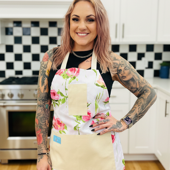 Woman wearing Amanda Bright Designs Vivien floral apron in a kitchen with white cabinets and black and white checkered tiles.
