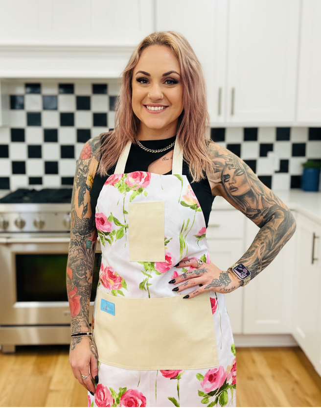 Woman wearing Amanda Bright Designs Vivien floral apron in a kitchen with white cabinets and black and white checkered tiles.