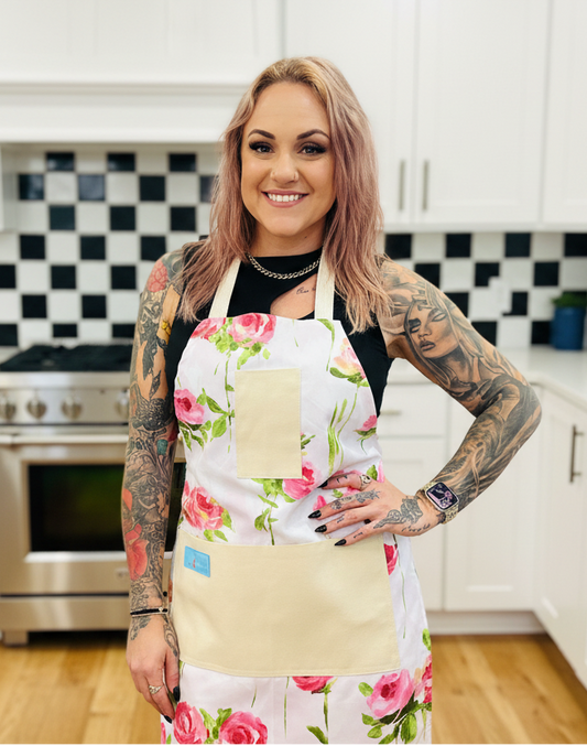 Woman wearing Amanda Bright Designs Vivien floral apron in a kitchen with white cabinets and black and white checkered tiles.