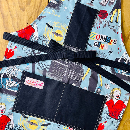 Amanda Bright Designs Halloween-horror themed apron with an aqua blue background and a zombie girl graphic print, featuring black fabric pockets and a black waist tie, with haunted houses, skeletons in coffins, a screaming woman in red, bats, full moon, black cats, blood dripping from roses and gravestones