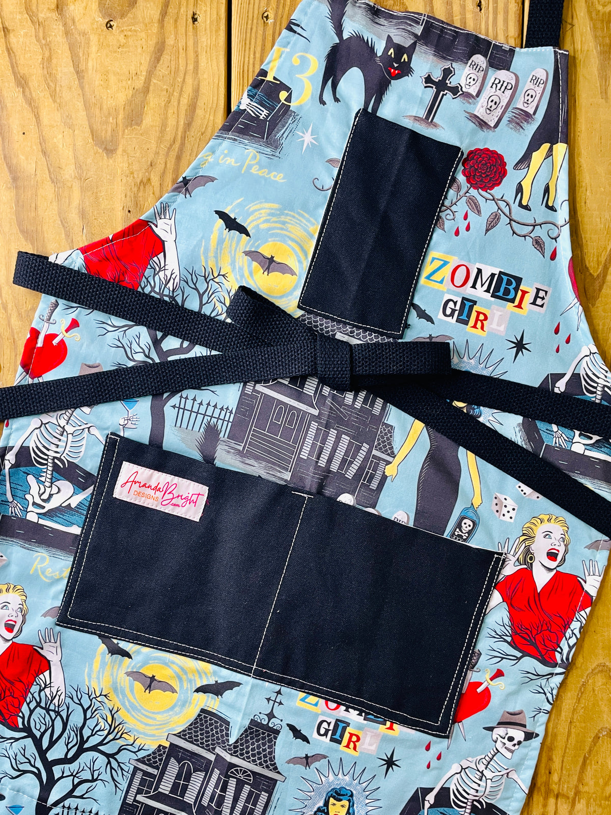 Amanda Bright Designs Halloween-horror themed apron with an aqua blue background and a zombie girl graphic print, featuring black fabric pockets and a black waist tie, with haunted houses, skeletons in coffins, a screaming woman in red, bats, full moon, black cats, blood dripping from roses and gravestones