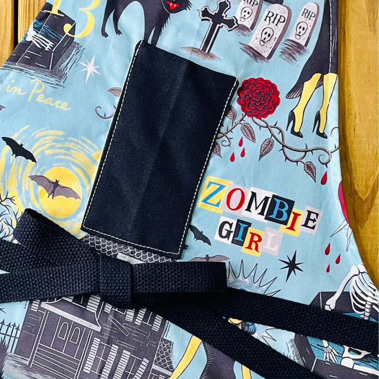 Amanda Bright Designs Halloween-horror themed apron with an aqua blue background and a zombie girl graphic print, featuring black fabric pockets and a black waist tie, with haunted houses, skeletons in coffins, a screaming woman in red, bats, full moon, black cats, blood dripping from roses and gravestones