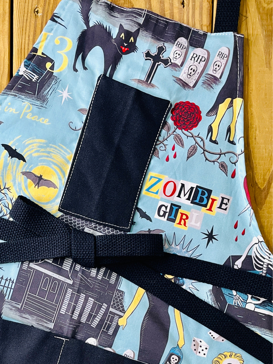 Amanda Bright Designs Halloween-horror themed apron with an aqua blue background and a zombie girl graphic print, featuring black fabric pockets and a black waist tie, with haunted houses, skeletons in coffins, a screaming woman in red, bats, full moon, black cats, blood dripping from roses and gravestones