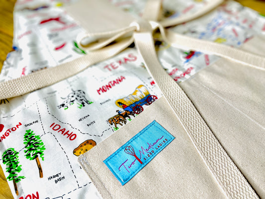 Two Medium Sized Ladies Sadie kitchen apron 50 states and their capitals historic industries road trip American heartland travel Conestoga wagon oil well longhorn airplane canoe cheese log cabin closeup view of apron